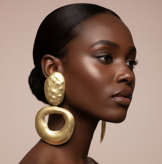 A woman wearing A pair of chunky gold-tone statement earrings with one oval textured drop and one larger open circular drop, displayed against a nude wall