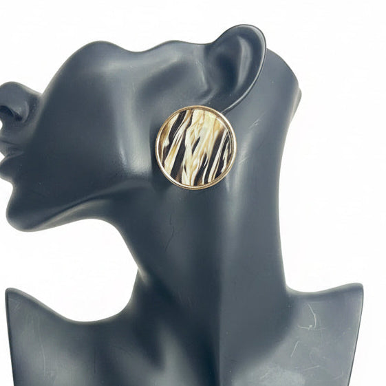 A single round gold-toned earring with a black and white zebra-like pattern, displayed on a matte dark gray female bust against a white background .