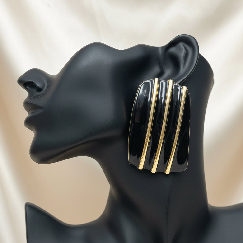 A black matte female bust sculpture displaying a pair of chunky black and gold striped dangle earrings, set against a champagne satin background