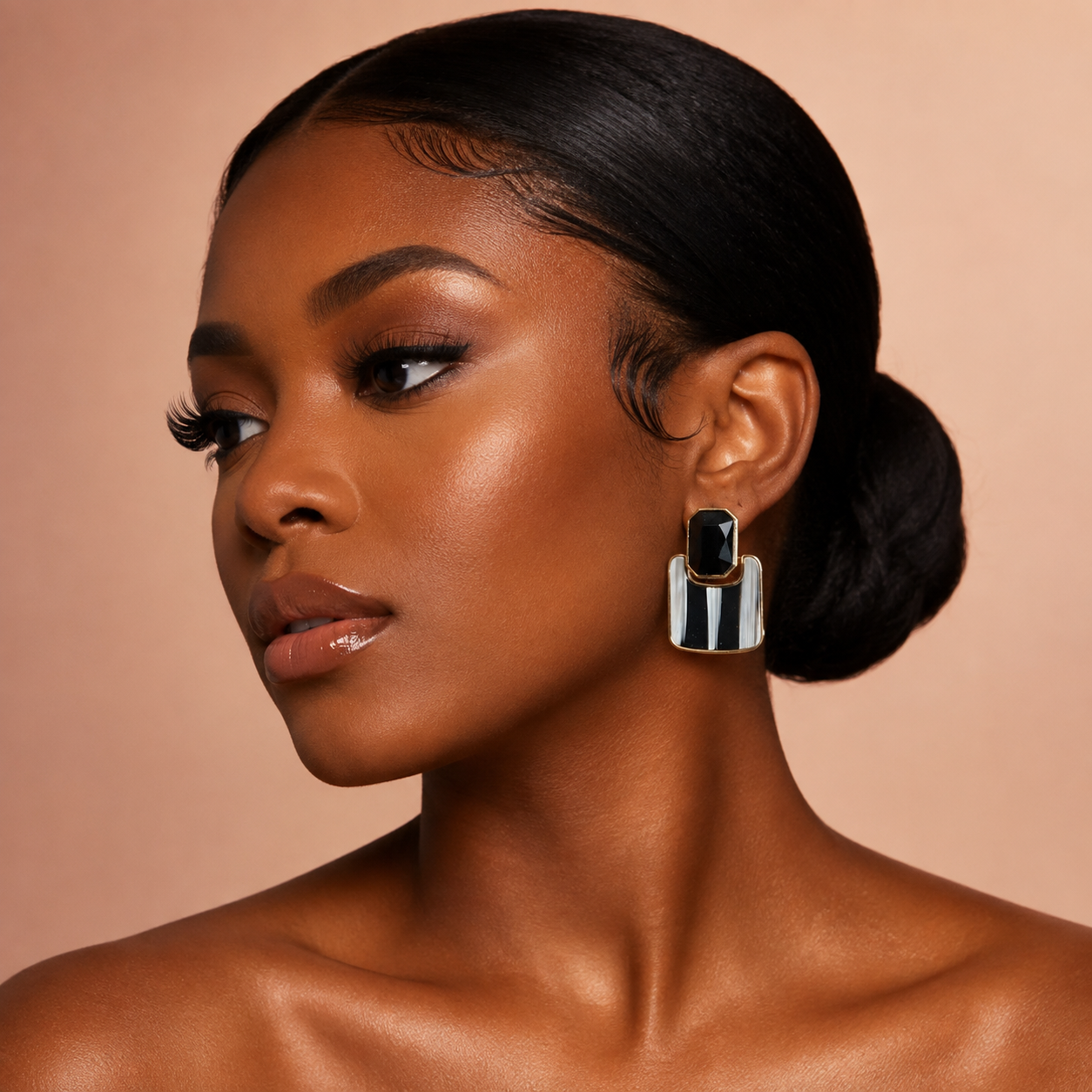 A close-up portrait of a woman with dark hair in a low bun wearing a bold geometric drop earring with black and silver panels, set against a solid warm beige background.
