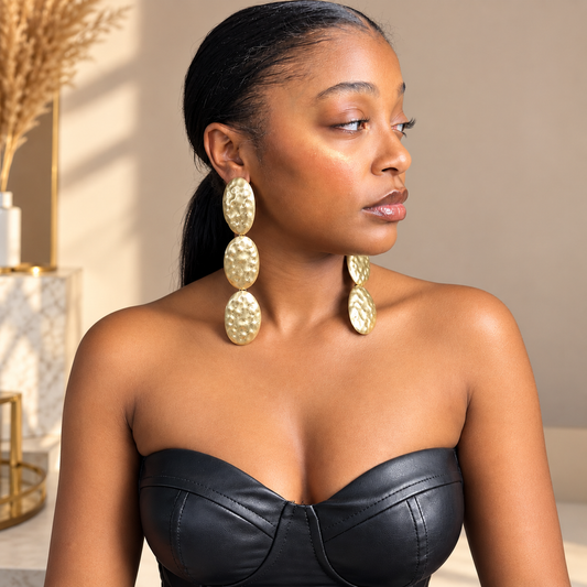 A woman wearing large, textured gold drop earrings with three stacked oval discs, paired with a black strapless top, shown in a warm indoor setting.