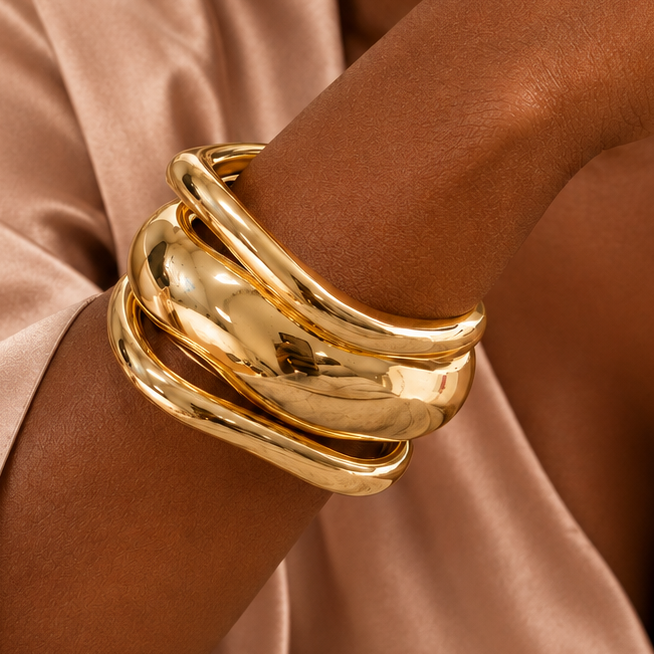 A close-up of a person wearing a stack of wide, polished gold bangle bracelets on their wrist, dressed in a satin champagne robe with a vase of dried flowers in the background.