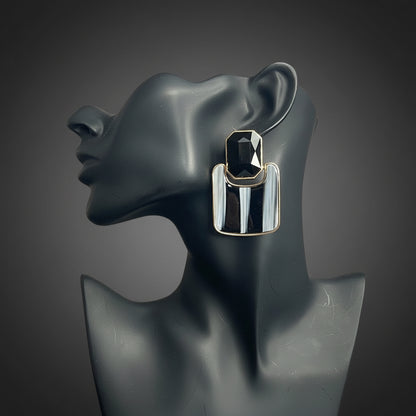 A pair of chunky silver-toned geometric drop earrings with black and white vertical stripes, displayed on a dark gray mannequin bust against a gray subway tile wall.