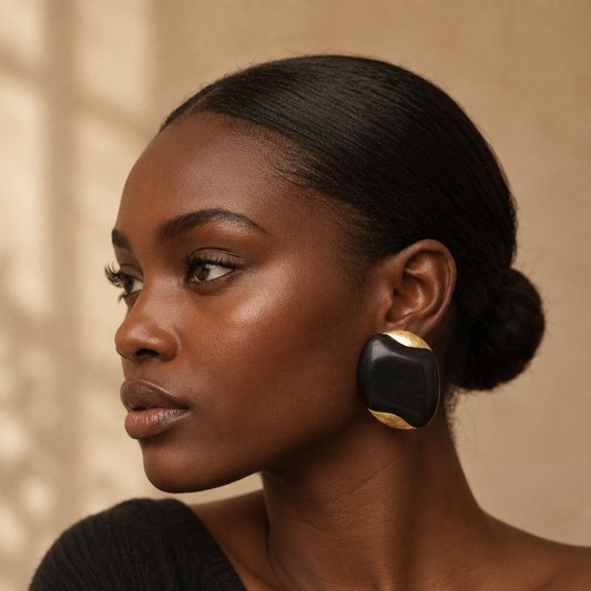 A close-up side profile of a woman with dark hair in a bun, wearing a large black and gold statement earring, set against a solid warm beige background.