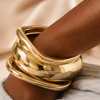 A close-up of a person's wrist wearing a stack of wide, polished gold bangles, resting on a light gray knit sleeve against a soft beige satin fabric background.
