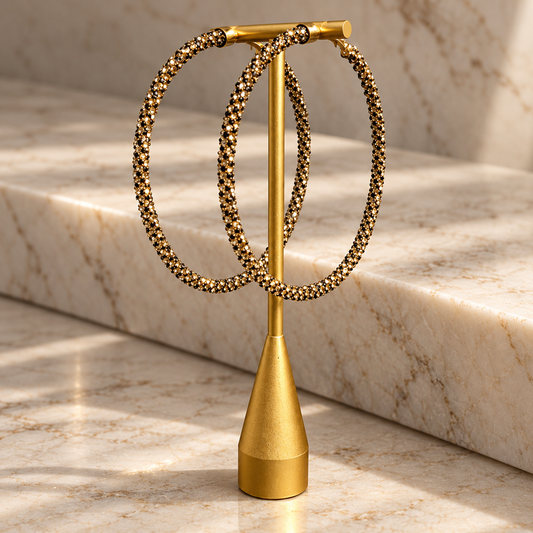 A pair of large gold hoop earrings with textured beaded edges, displayed on a gold stand against a marble background.