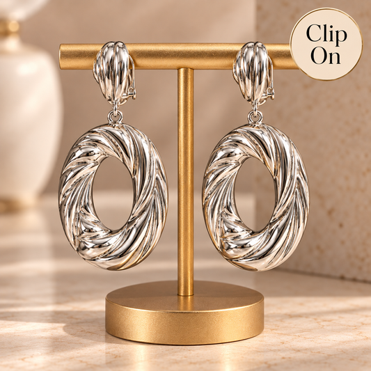 A pair of chunky silver hoop earrings with a twisted rope texture, hanging from matching silver clip-on hooks from a gold jewelry display stand. A beige circular badge in the top right corner reads 'Clip On'.