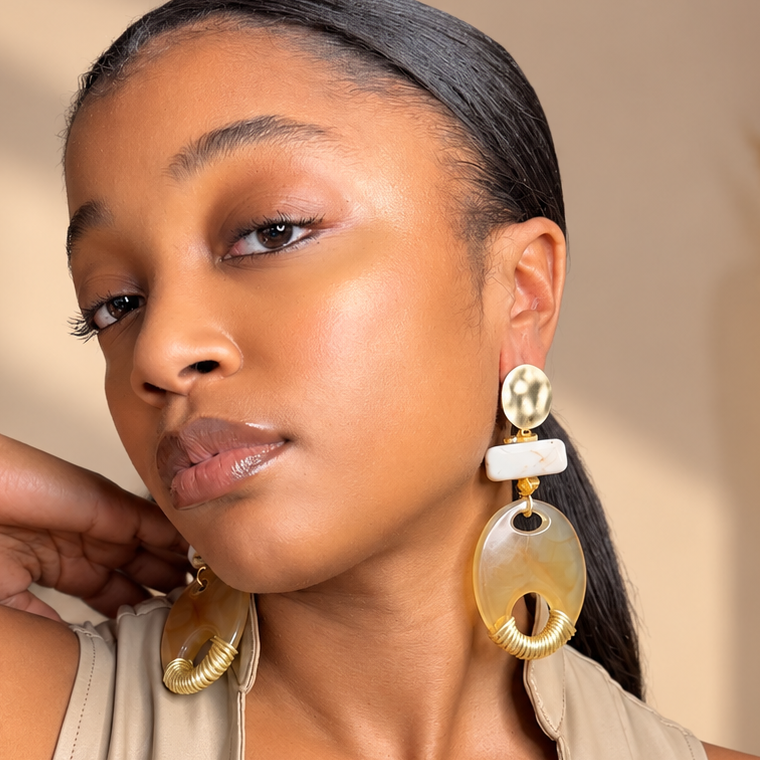 A woman wearing large statement earrings with oval amber and white stone elements, paired with a beige lace-up top in a warm indoor setting.
