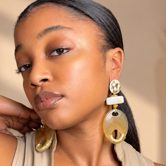 A woman wearing large statement earrings with oval amber and white stone elements, paired with a beige lace-up top in a warm indoor setting.
