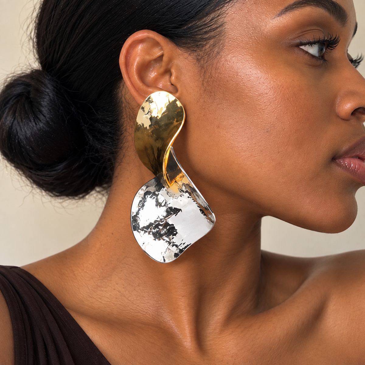 A close-up of a woman wearing a large, sculptural statement earring. The earring features a polished gold-toned top section and a textured, hammered silver-toned bottom section 