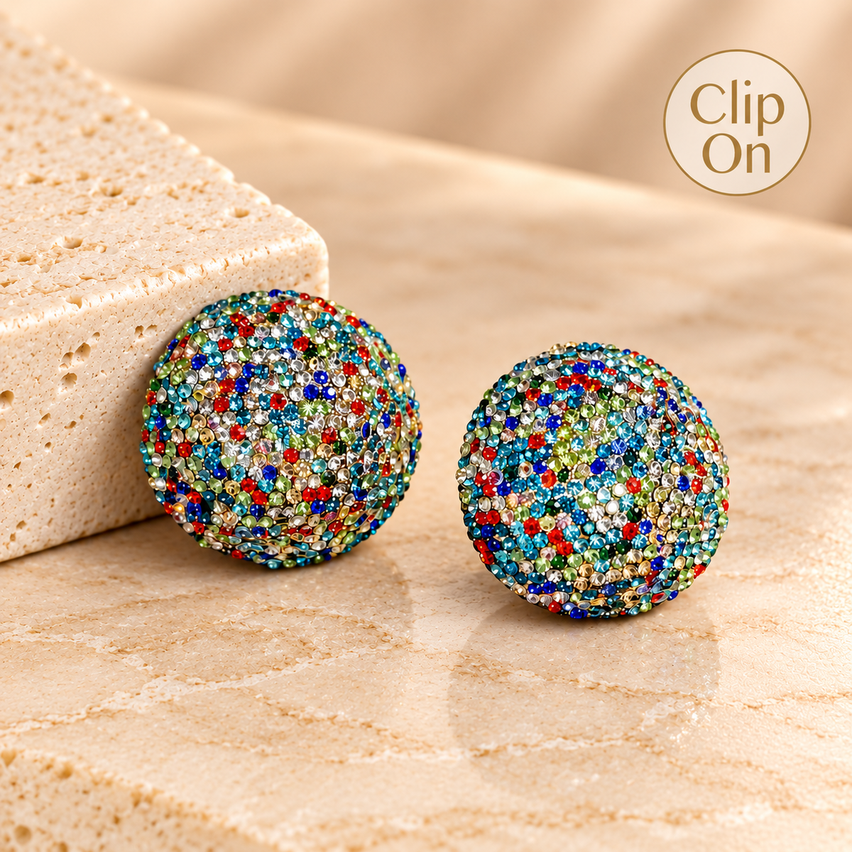 A pair of round stud earrings covered in dense multicolor rhinestones, displayed on a beige stone surface next to a textured stone block. A 'Clip On' badge is visible in the top right corner.