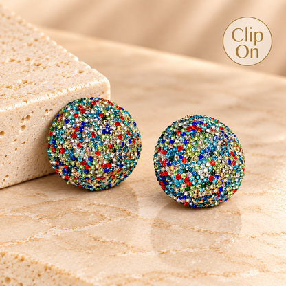 A pair of round stud earrings covered in dense multicolor rhinestones, displayed on a beige stone surface next to a textured stone block. A 'Clip On' badge is visible in the top right corner.