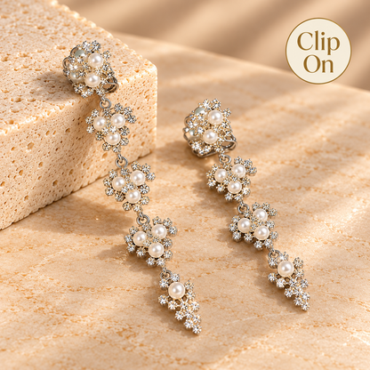 A pair of silver-toned drop earrings with clusters of white faux pearls and clear rhinestones, displayed on a textured beige surface next to a stone block, with a 'Clip On' badge in the top right corner.