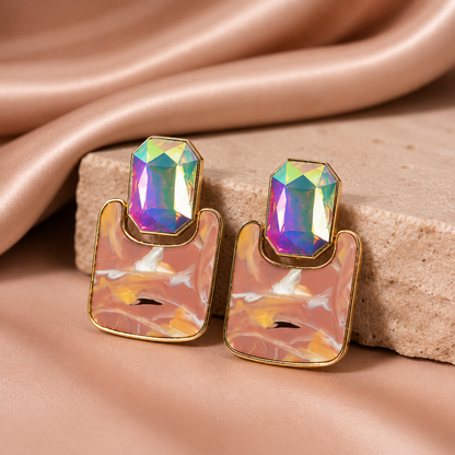 A pair of gold-tone statement earrings with iridescent, rainbow-flecked rectangular gemstones on top of blush pink marbled resin, displayed on a beige stone block with draped satin fabric.