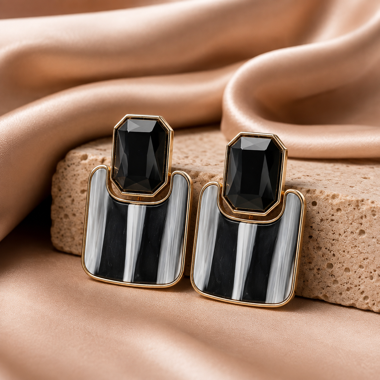 A pair of geometric statement earrings with faceted black stones and black-and-white striped inlay, set in gold-tone metal, displayed on a textured stone slab with draped beige satin fabric.