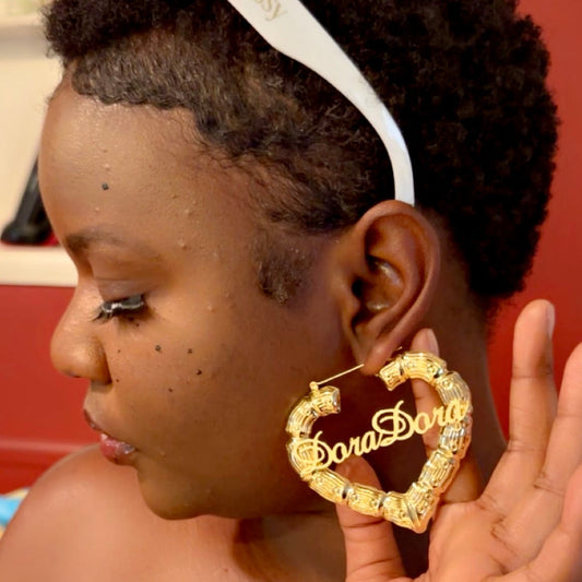 Close-up side profile of a woman wearing a large gold bamboo-style heart hoop earring with a personalized nameplate reading “DoraDora” holding the earring to highlight its size and shine.